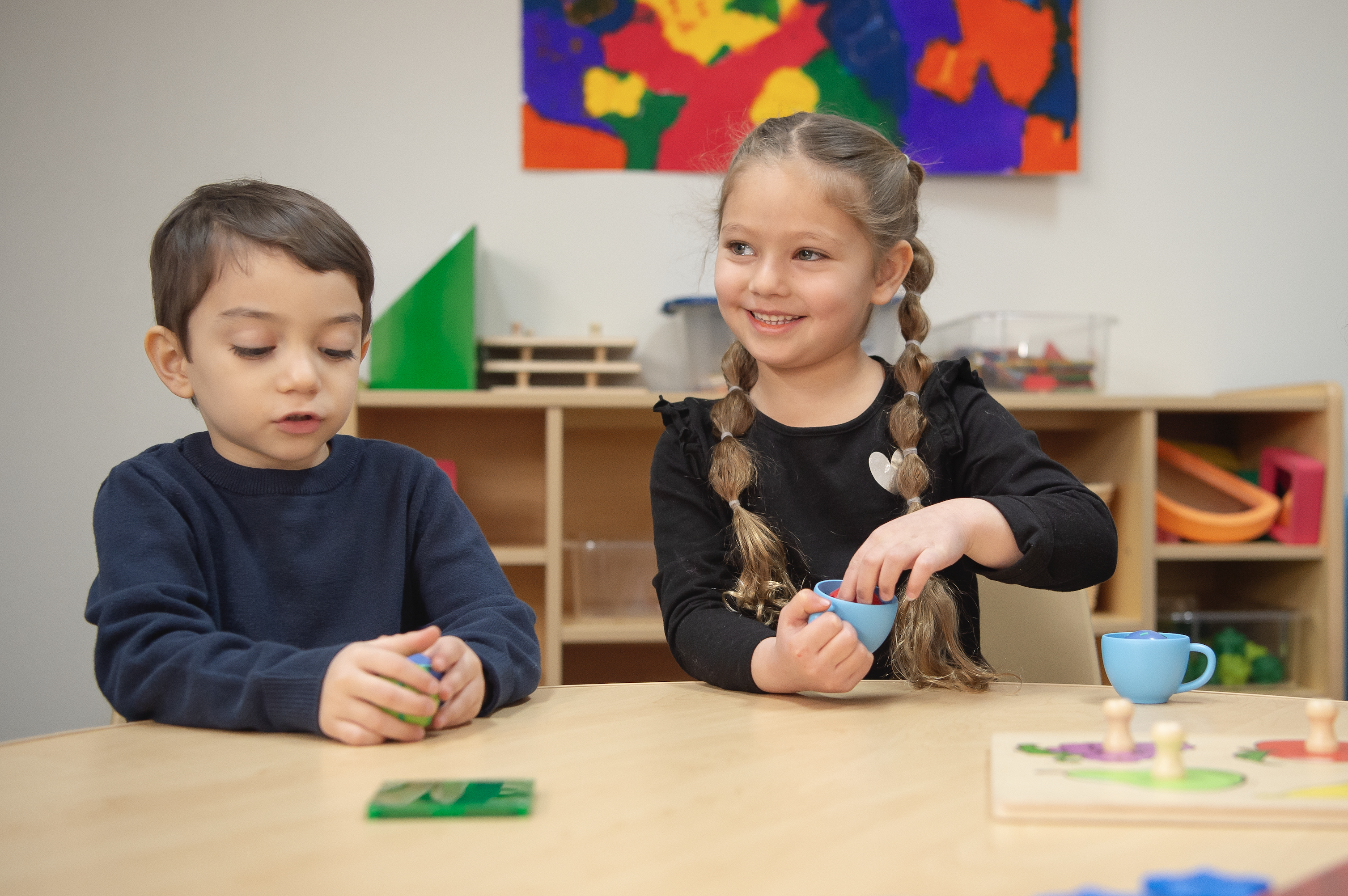 Two toddlers sitting at a table in a classroom setting, smiling playing with toys on the table. 