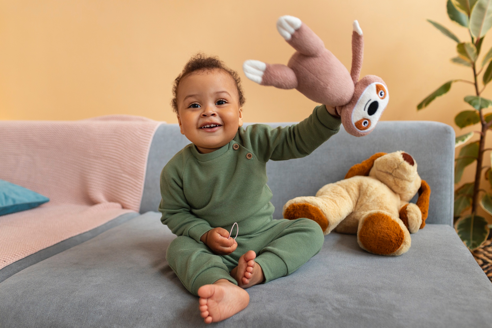 Smiling toddler wearing dark green onesie sits on a couch holding up a stuffed animal and smiling.