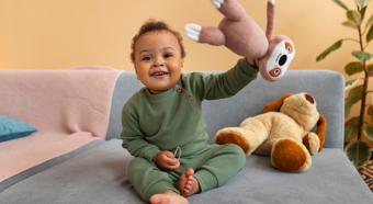 Smiling toddler wearing dark green onesie sits on a couch holding up a stuffed animal and smiling.