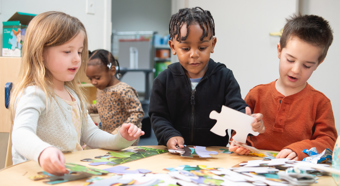 Three toddlers working on a puzzle at a table in a classroom setting. 