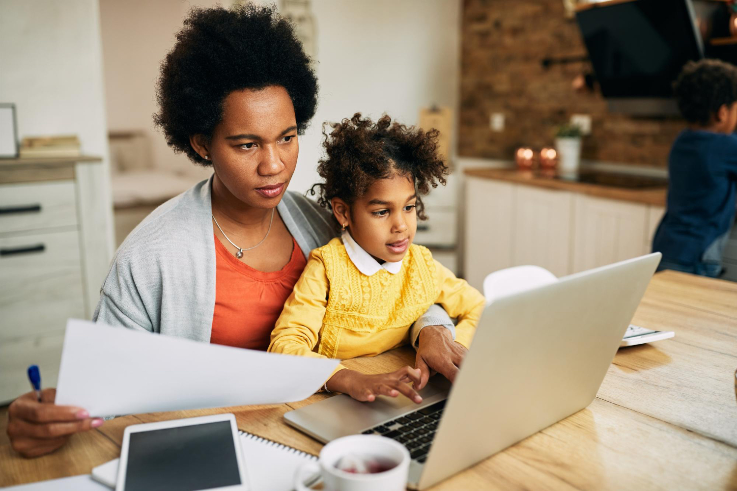 Adult female sitting at a table with toddler in her lap while looking at a computer screen and papers in her hands.