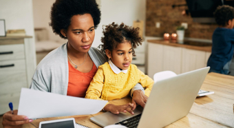 Adult female sitting at a table with toddler in her lap while looking at a computer screen and papers in her hands.