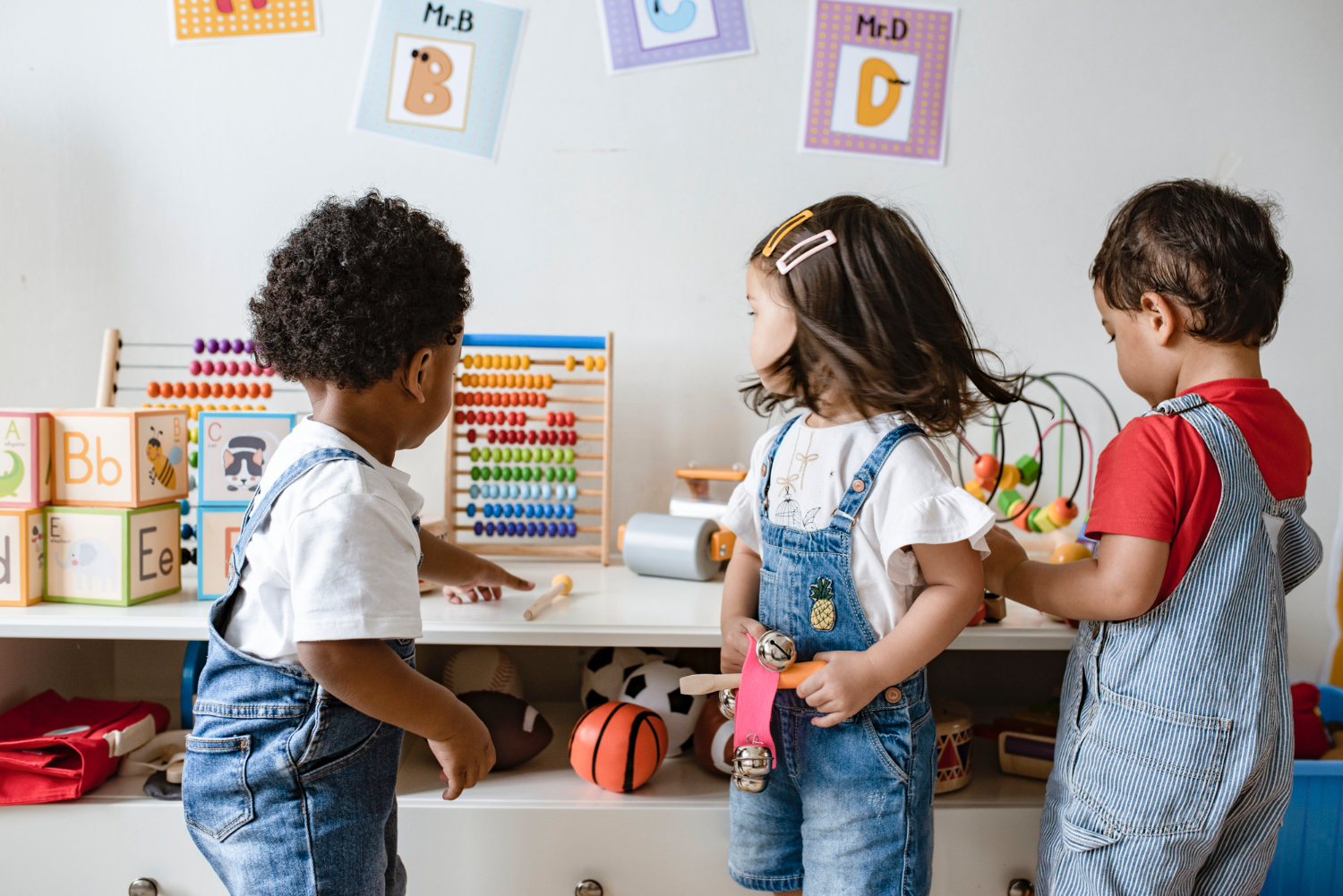 Tres niños pequeños, todos ellos vestidos con overoles vaqueros, juegan juntos con juguetes en un aula.