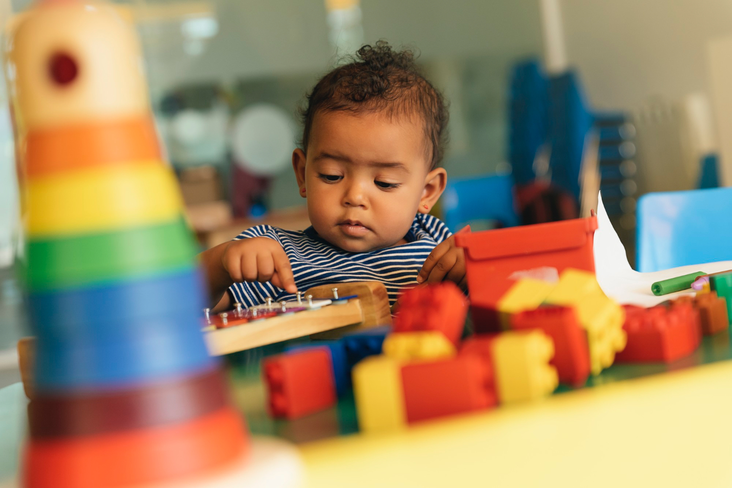 Toddler sitting at table in colorful play setting, toy blocks and other toys on the table. 