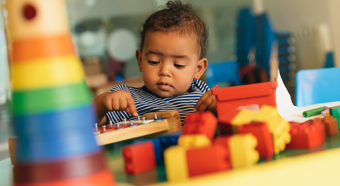 Toddler sitting at table in colorful play setting, toy blocks and other toys on the table. 